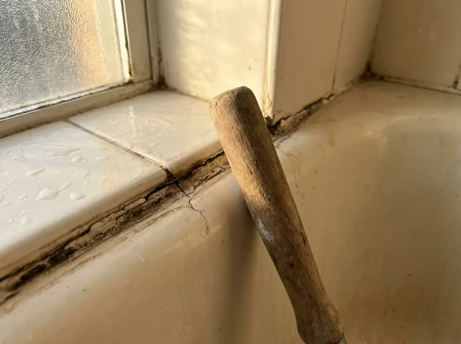 Joint de baignoire noirci près d’une fenêtre, outil en bois posé contre le carrelage